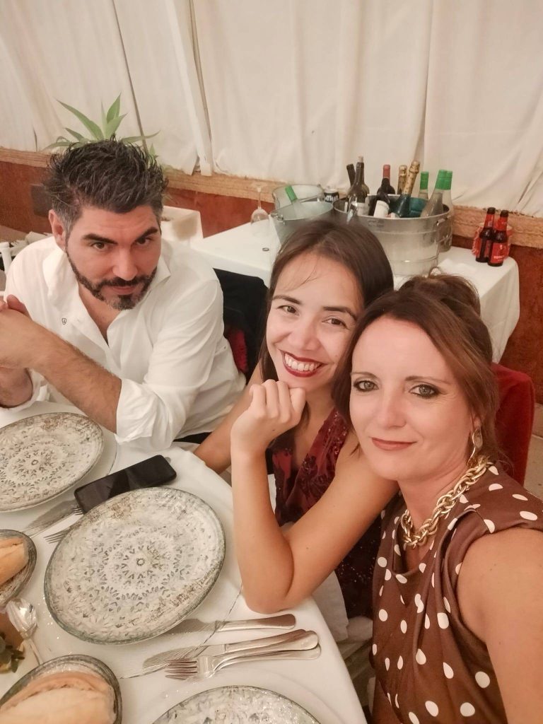Angel, Shayna, and Raquel smiling together at a dinner table set with elegant plates and glassware, at a Spanish family wedding.